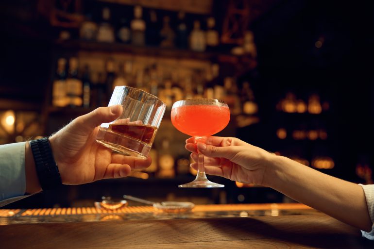 Man en vrouw proosten hun drankjes met elkaar. Een bar staat op de achtergrond.