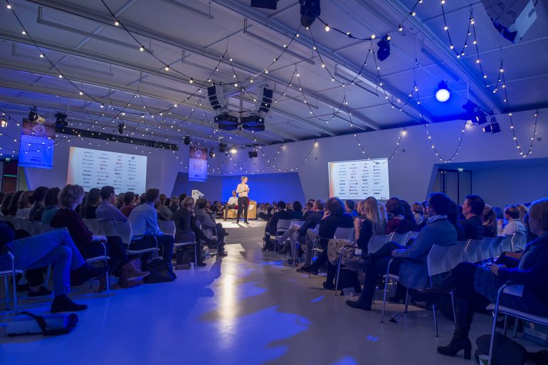 Zaal in grote ruimte kijkt naar spreker op podium